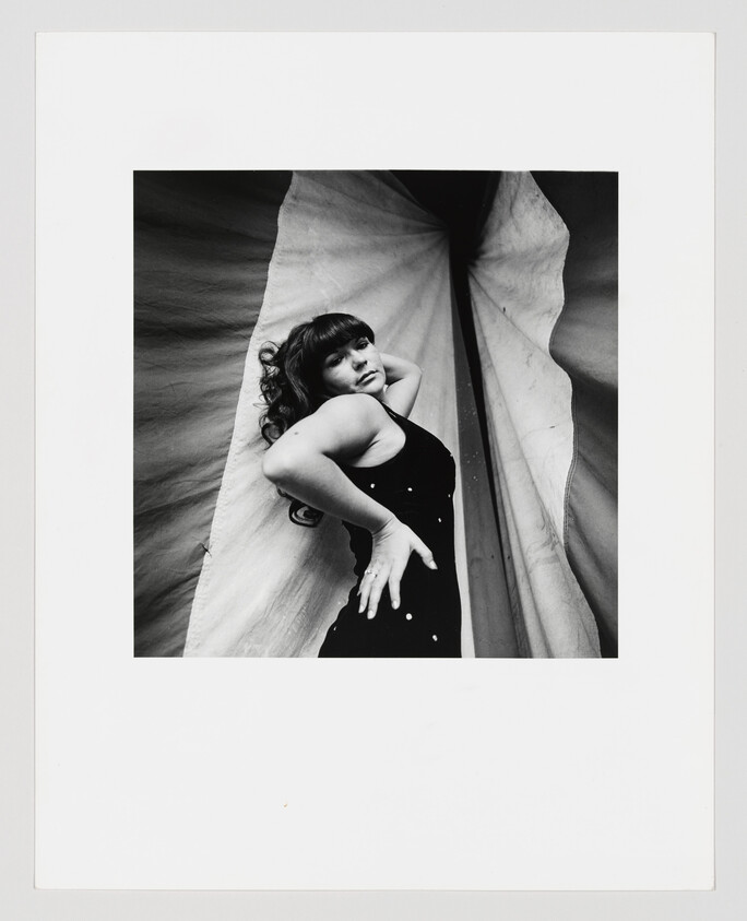 A woman in a black dress poses with one hand on her hip against draped fabric.