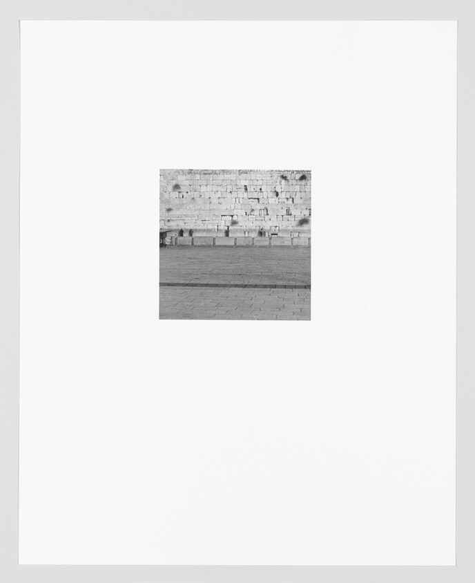 A black and white photograph centered on a section of an ancient stone wall with visible layers of large rectangular blocks. The wall is set against a clear sky, and there's a paved area in the foreground with no visible people. The image is framed with a wide white border.