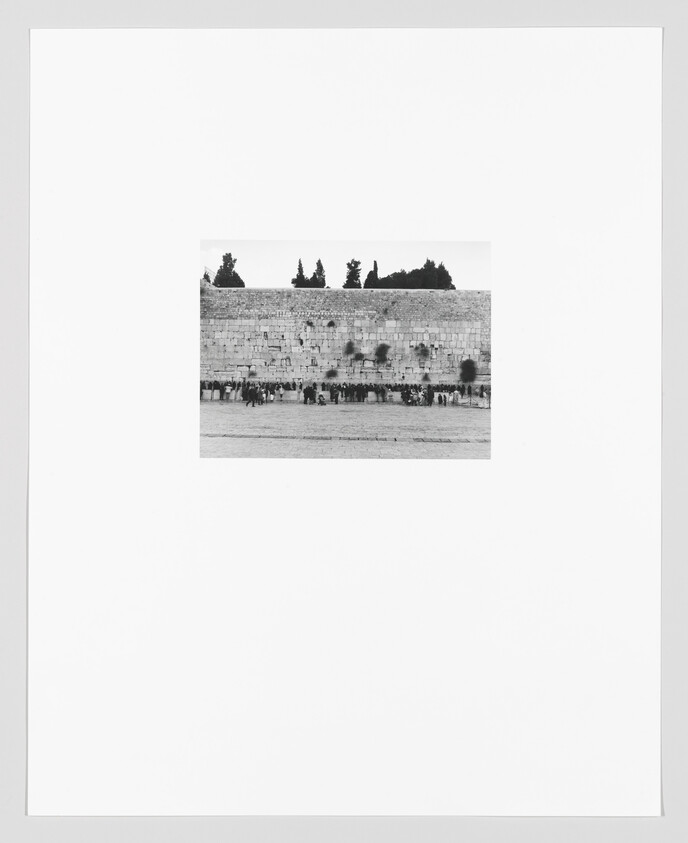 Black and white photograph of the Western Wall in Jerusalem with several people gathered in front of it. The wall is shown with a clear sky above and trees peeking out from the top edge.
