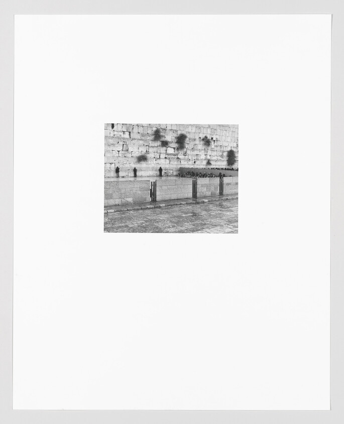 Black and white photograph of the Western Wall in Jerusalem with several people standing in front of it, centered within a large white border.