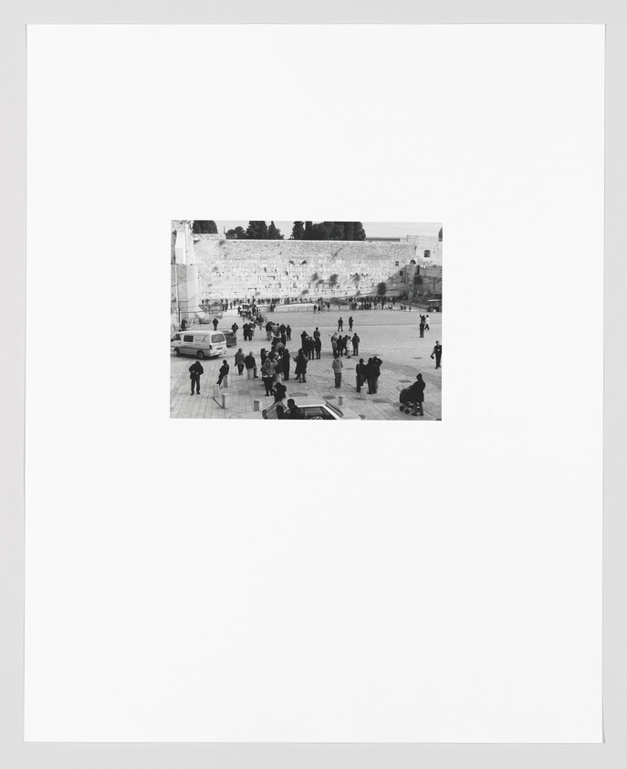 Black and white photo of visitors at the Western Wall in Jerusalem, with people standing and walking in the plaza in front of the ancient stone wall.