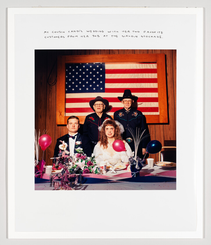 Bride and groom sit at decorated table with two men in cowboy hats and a large American flag.