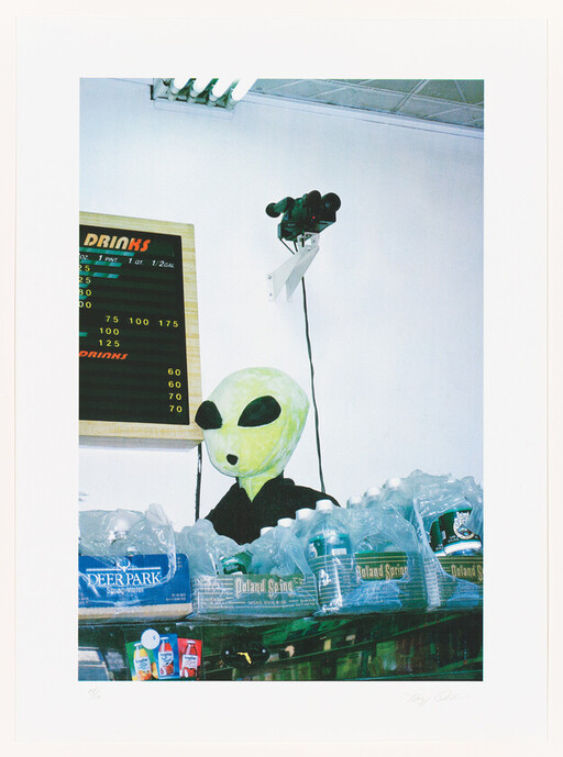 A green alien mannequin head stands behind packs of bottled water in a store.