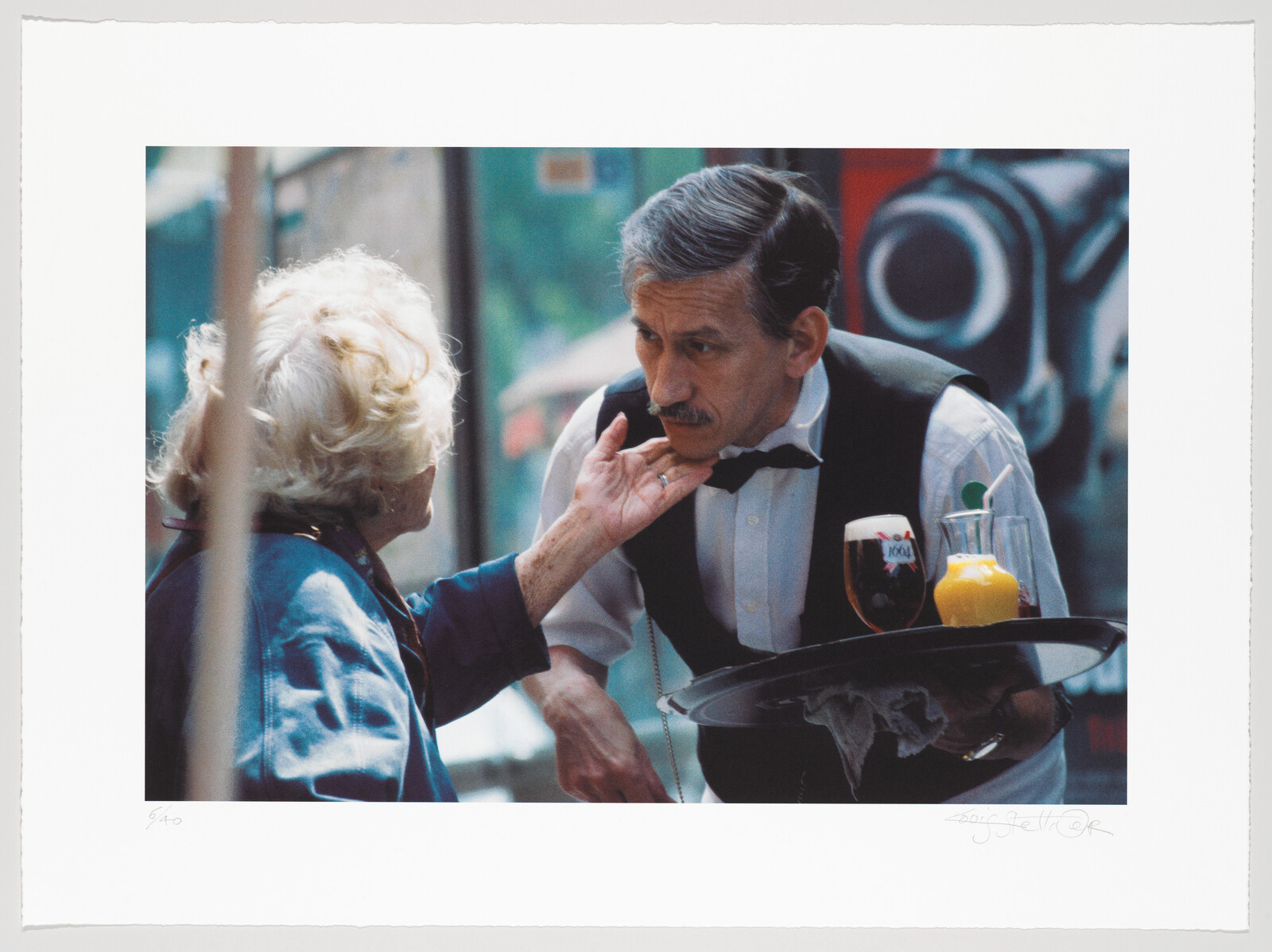 An elderly woman gently touches a waiter’s chin while he leans forward holding a tray of drinks.