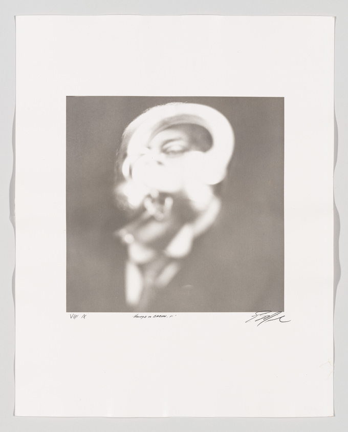 A blurry black and white photograph of a person with exaggerated facial features, possibly singing or shouting, framed within a larger white border that includes handwritten text and a signature at the bottom.