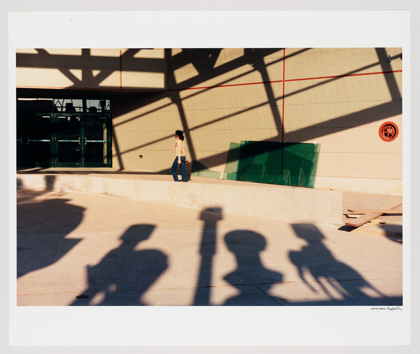 A person walks along a raised concrete ledge under strong geometric shadows while onlookers cast long silhouettes.