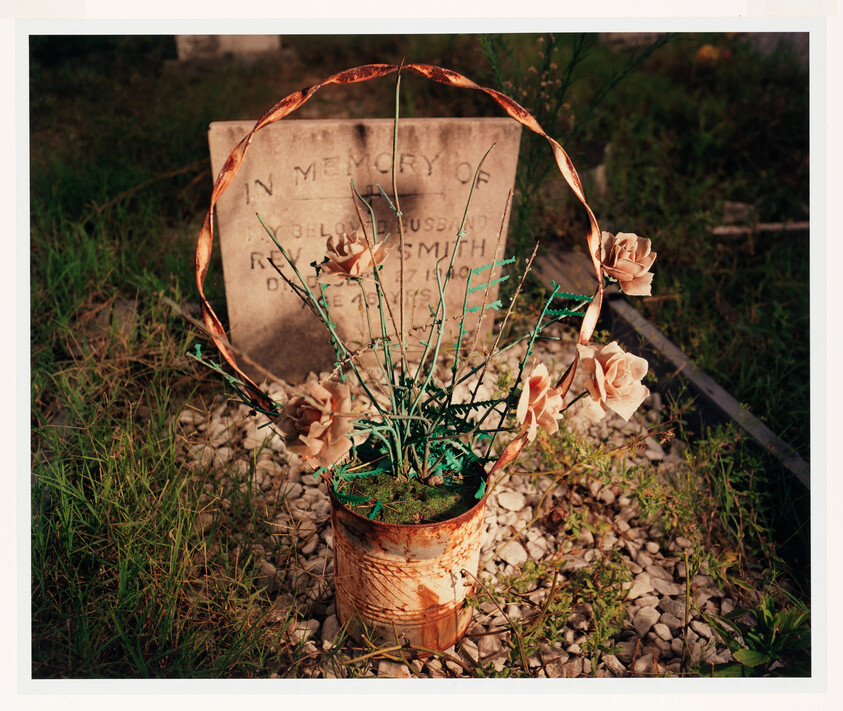 A rusty metal bucket holds artificial roses and greenery placed at a gravesite.
