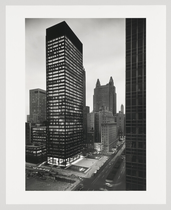 Tall glass skyscraper towers over a lit plaza and busy city street at dusk.