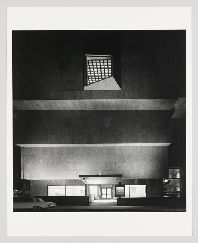 Black and white photo of a modern building at night with geometric shapes and a vintage car parked outside.