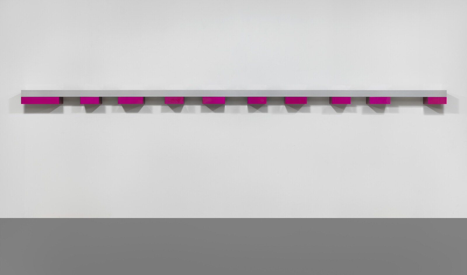 A long narrow white shelf mounted on a wall with evenly spaced bright magenta blocks underneath.