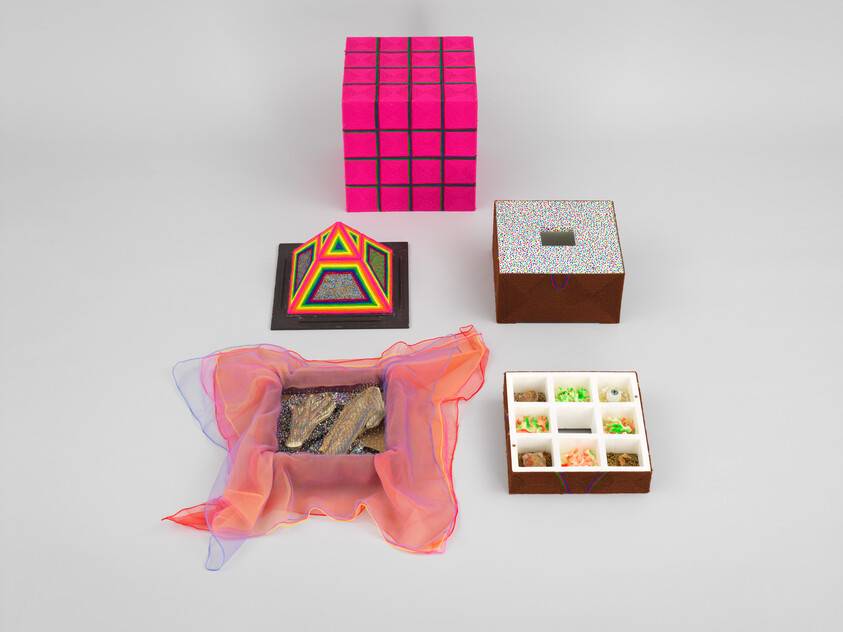 Five small colorful boxes and a pink cloth arranged on a gray surface, including a neon pyramid and pink cube.
