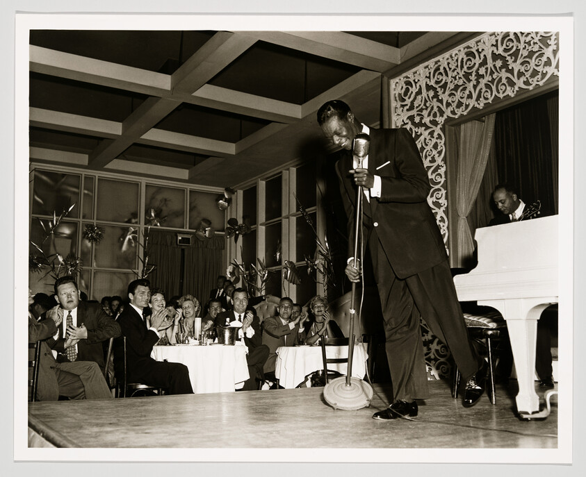 Tall singer in suit leans into microphone while an applauding audience watches and a pianist accompanies.
