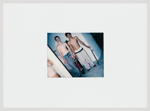 Two shirtless teenage boys stand indoors holding skateboards, one leaning against a wall.