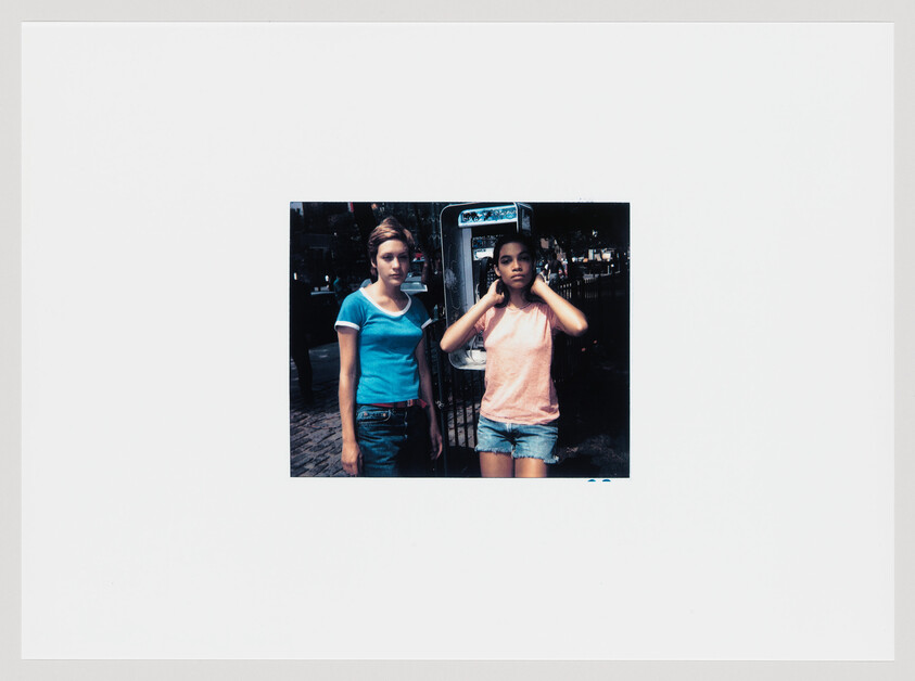 Two teenage girls stand by a public phone, one adjusting her hair and the other watching.