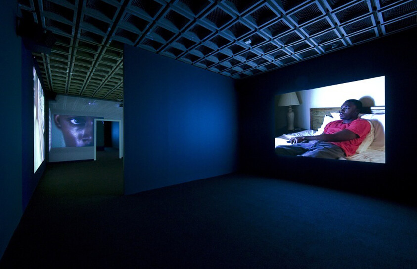 Dark gallery room with two large video projections, one showing a man reclining on a bed.