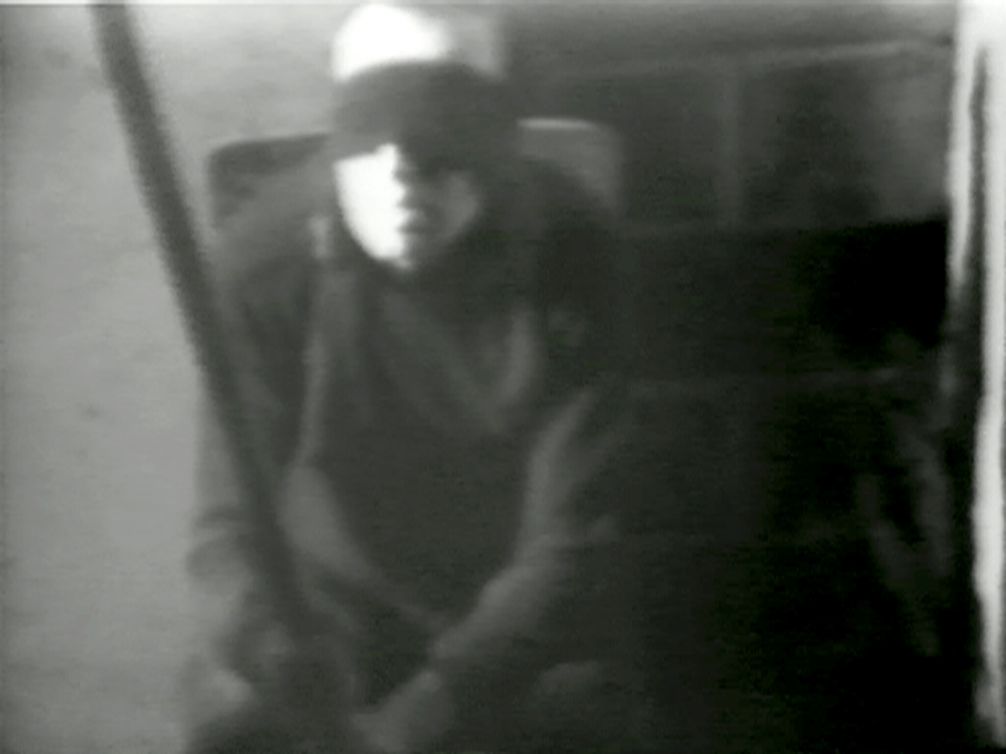 A grainy black-and-white scene shows a person wearing a cap sitting with face partially illuminated.