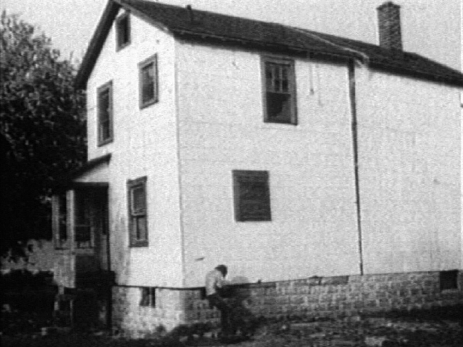 Two-story white house with a person sitting by the stone foundation near the corner.