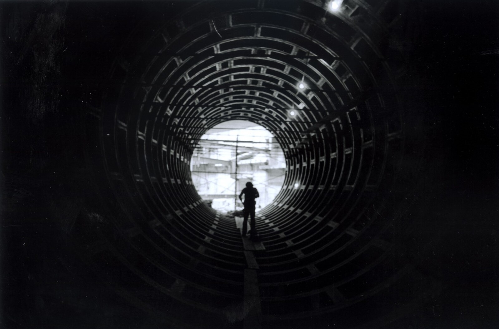 A worker walks along a narrow plank inside a large circular tunnel toward a bright exit.