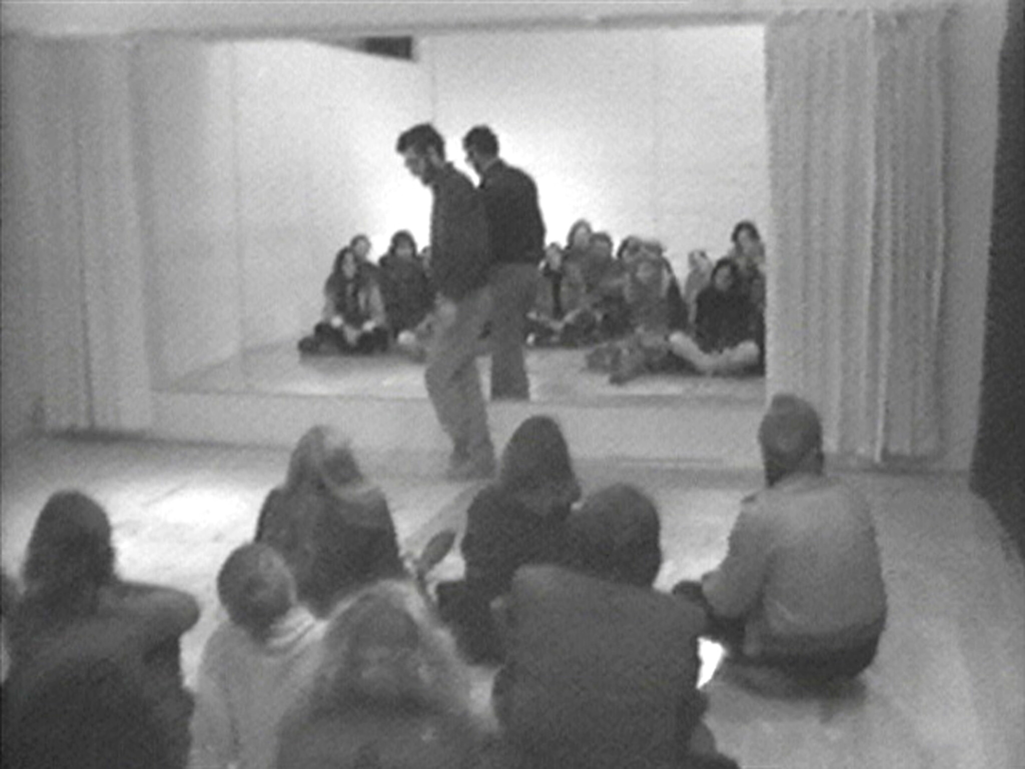 A group of people sit on the floor watching two people standing and moving in front of a large mirror.