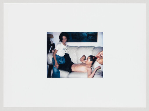 A woman sits on a couch while a shirtless man reclines and someone brushes his hair.