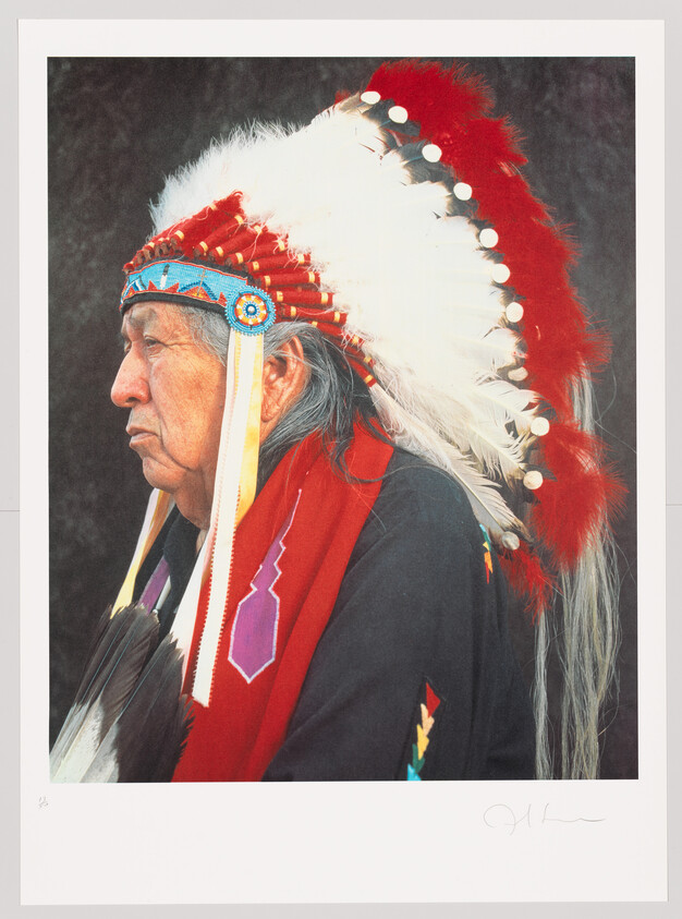 Profile of a Native American elder wearing a traditional feather headdress with red and white feathers and beadwork.
