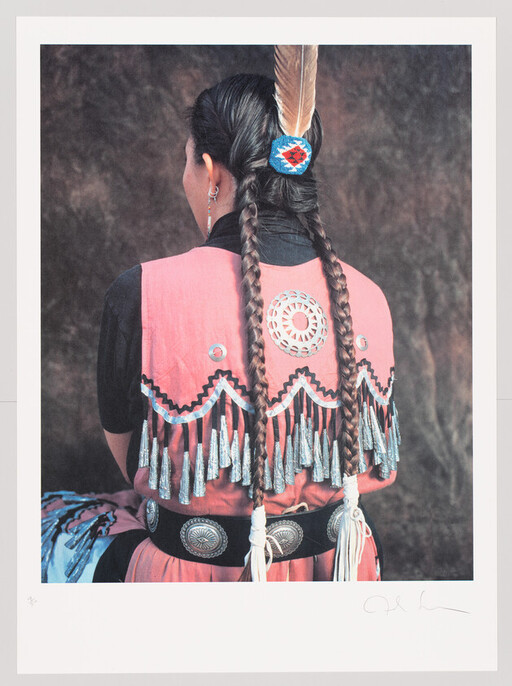 Woman with two long braids and a feather wears a pink fringed traditional dress.