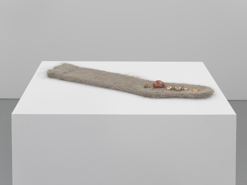 A long fuzzy fabric strip with several small beads and stones rests on a pedestal.