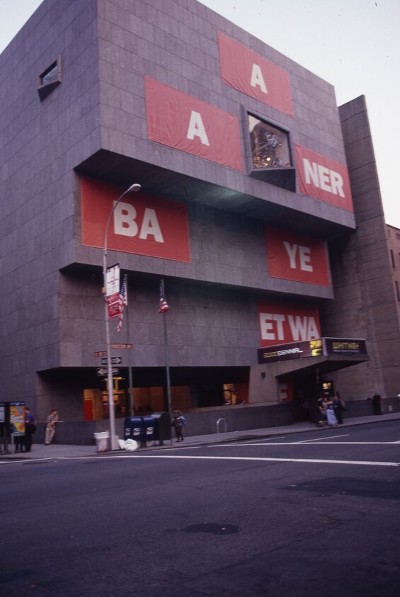Large gray building with red banners displaying partial white letters, people standing on the sidewalk, and two American flags.