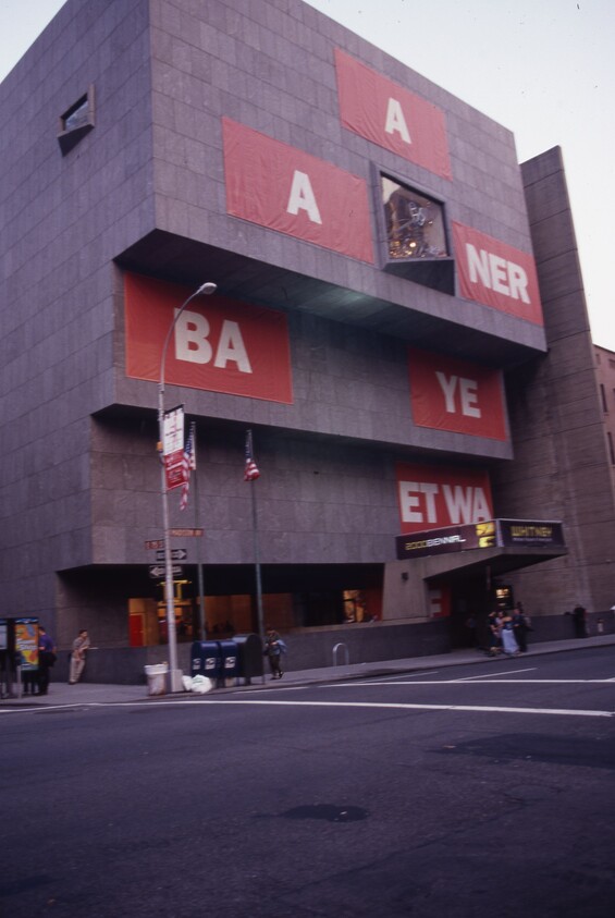 Brutalist museum building with large red banners displaying white letters and people entering the lobby.