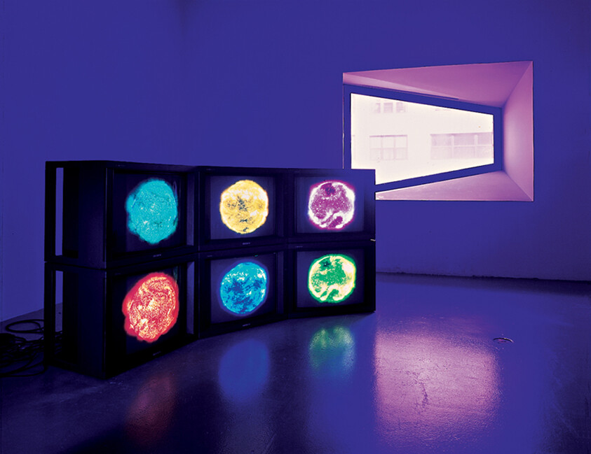 A contemporary art installation featuring a stack of six monitors in a dark room, each displaying a different brightly colored abstract image, with a purple hue illuminating the space and a large angled window casting daylight into the room.
