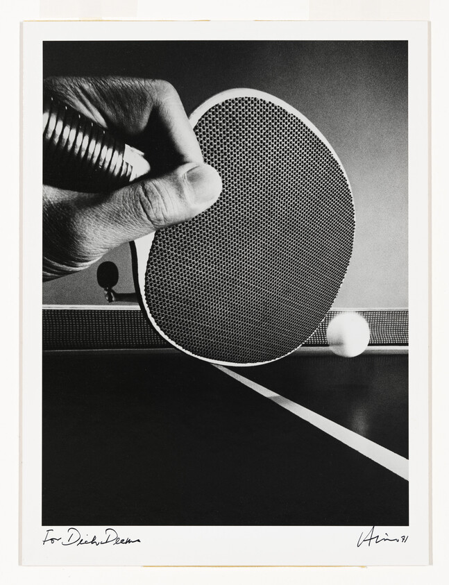 A hand holds a ping-pong paddle poised to hit a ball over the net on a table.