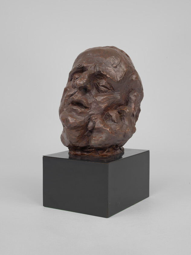 Bronze-textured sculpted human head with closed eyes and slightly open mouth resting on a black pedestal.