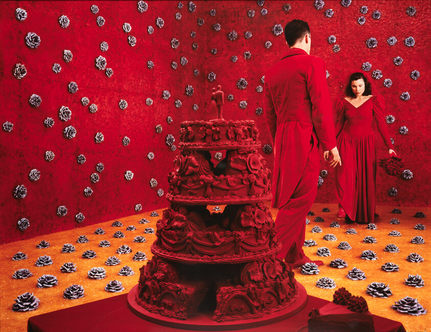 A large ornate red cake sits center while a man and woman in red face each other in decorated room.