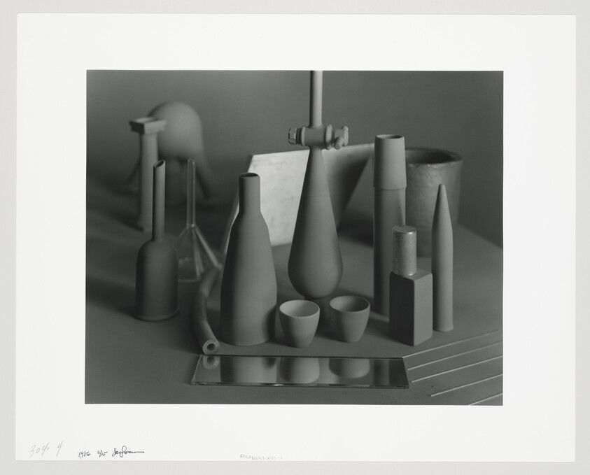 A still life arrangement of matte bottles, cones, cups, and a small mirror on a table.