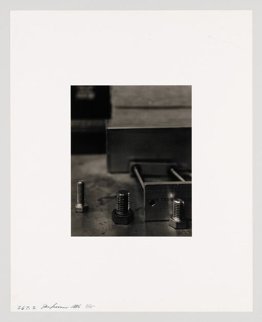 Three bolts and a metal block sit on a workshop table with two threaded rods.