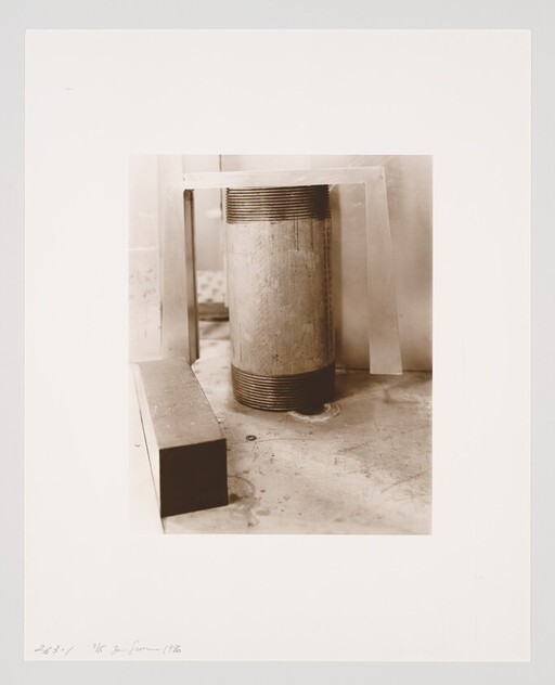 A vertical cylindrical pipe with threaded ends stands beside a rectangular block and metal braces.