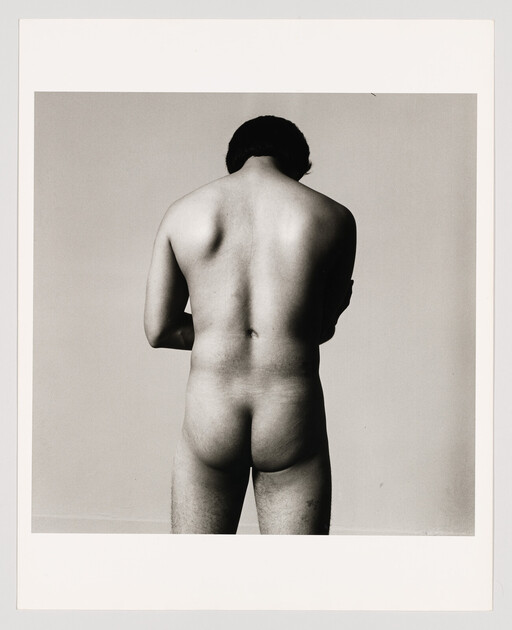 A person stands nude facing away, showing their bare back and buttocks against a plain background.
