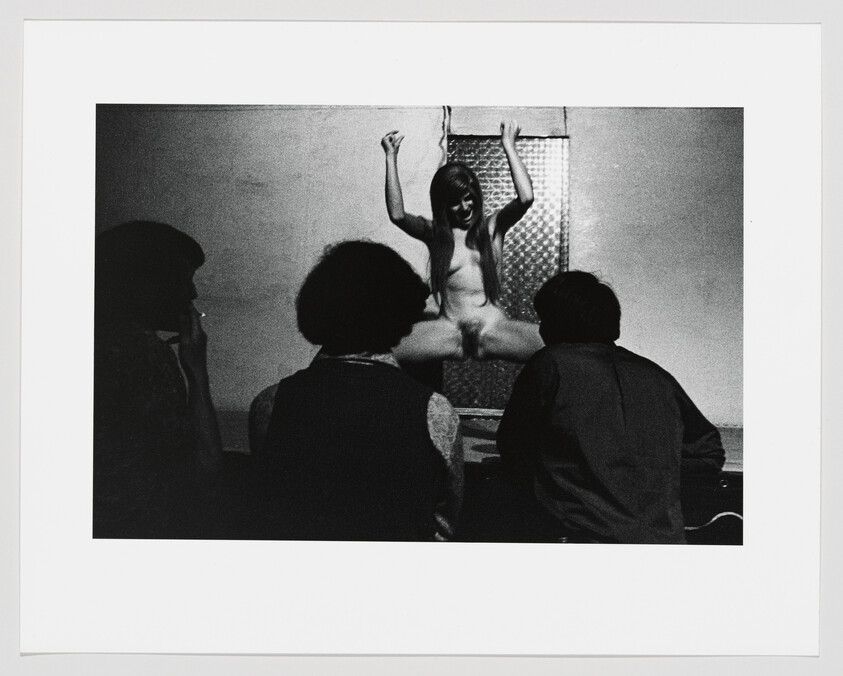 Young nude woman sits with legs spread on a ledge while three people watch from the foreground.