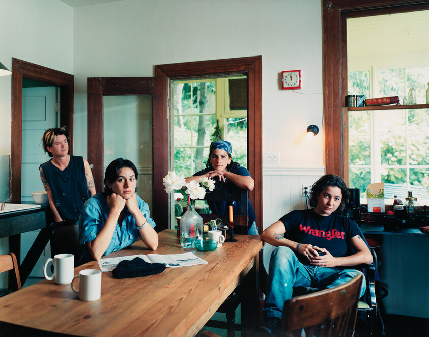 Four people sit around a kitchen table, looking toward the camera with mugs and a vase of flowers.