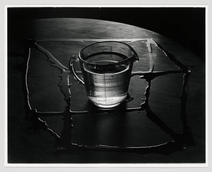 Glass measuring cup sits in the center of a dark table with spilled water forming a square.