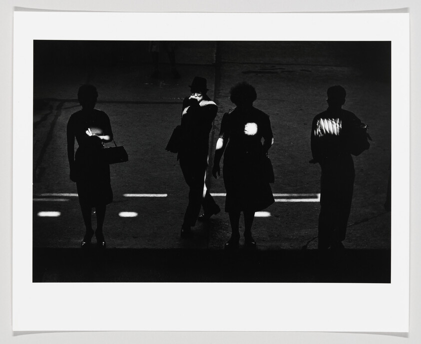 Four silhouetted pedestrians cross a dark street with bright patches of light on their bodies.