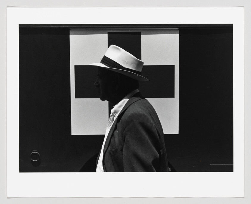 A black and white photograph featuring a profile view of a person wearing a white hat and a dark suit, standing in front of an abstract geometric background with a large black square and smaller white and black rectangles. The image has a strong contrast between the dark and light elements, and the person's silhouette is sharply defined against the patterned backdrop.
