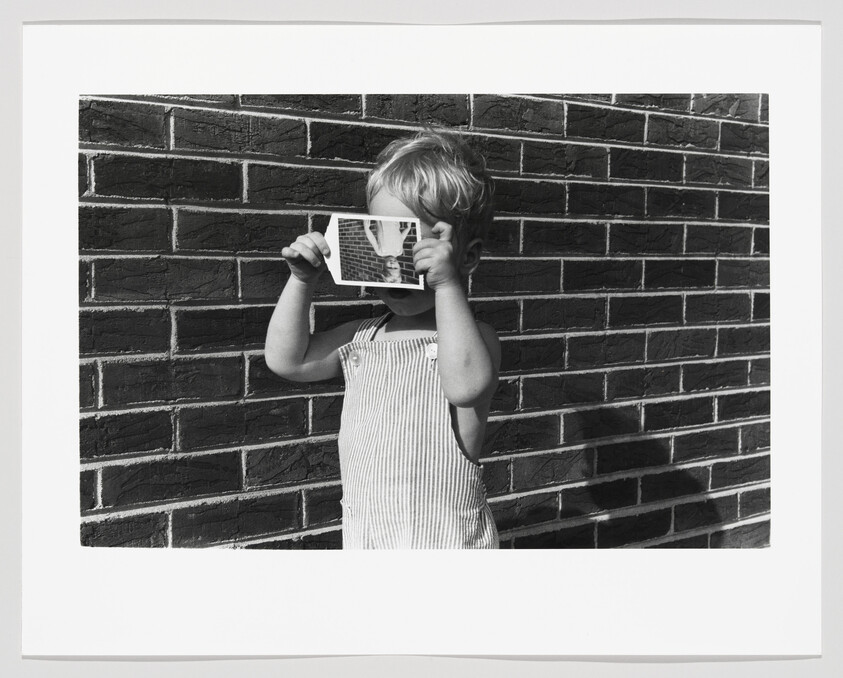Young child stands against a brick wall holding a small photograph up to their face.