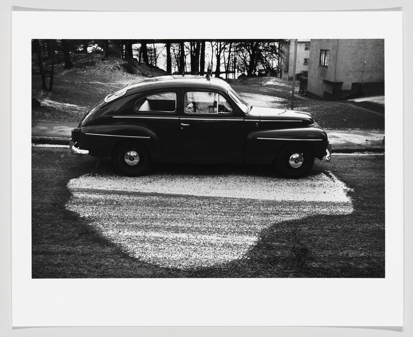A vintage dark car parked over a pale gravel patch on a quiet street.