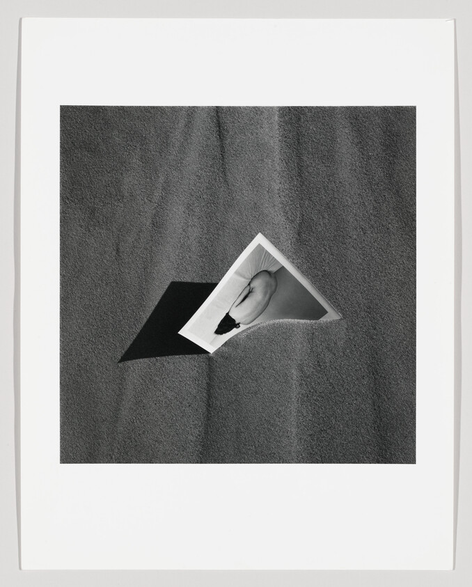 A black-and-white print of a reclining figure partly buried in textured sand casting a dark shadow.