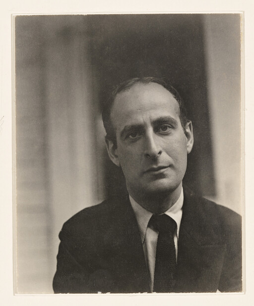 A solemn man in a dark suit and tie looks directly at the camera in a close portrait.