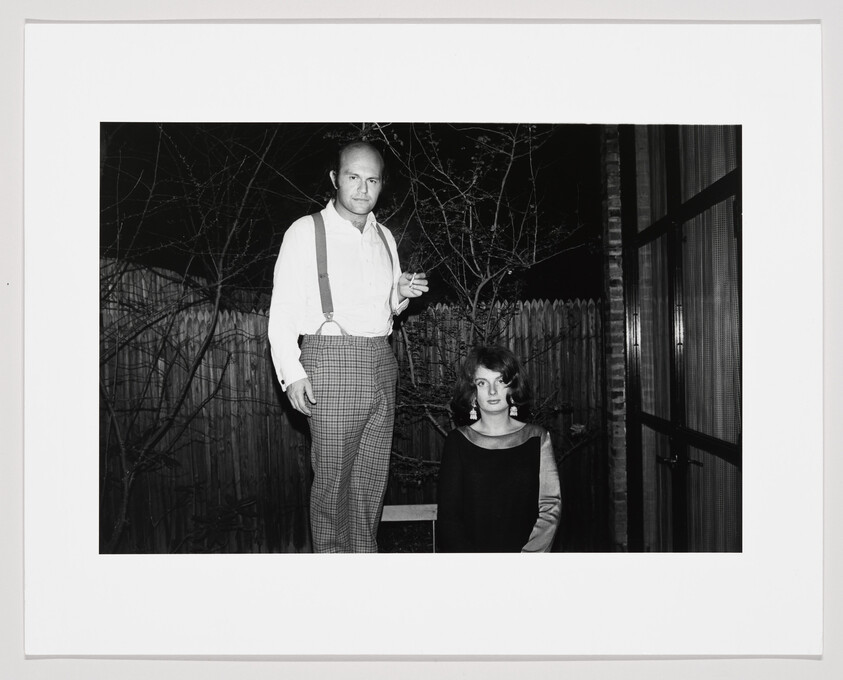 A black and white photograph featuring a man standing and a woman sitting in a night-time outdoor setting. The man is dressed in a white shirt with suspenders and checkered trousers, holding a cigarette, while the woman is wearing a dark top with a scoop neckline. They are positioned in front of a wooden fence and a glass door, with bare tree branches in the background.