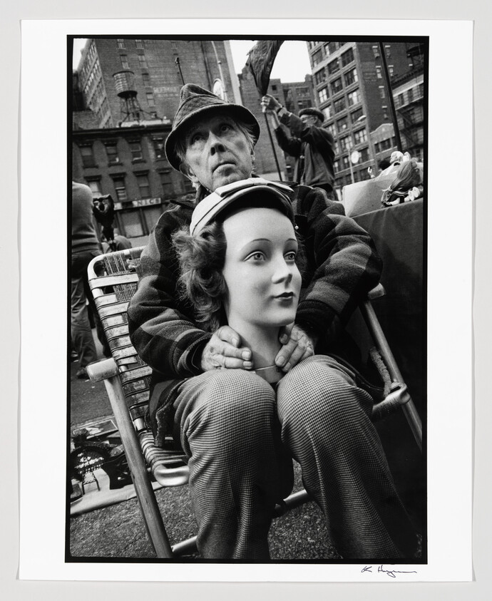 An older man sits at a street market cradling a mannequin head on his knees.