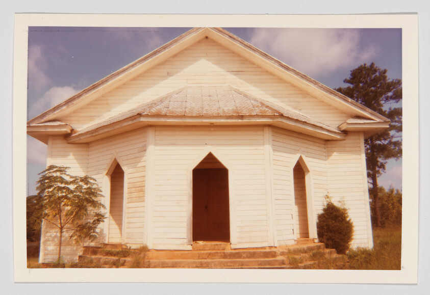 A vintage photograph of a small white wooden church with a gabled roof and a central entrance flanked by two narrow windows, set against a backdrop of trees and a blue sky with light clouds.