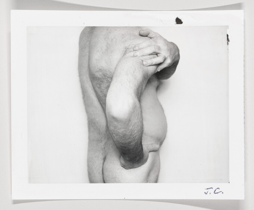 Black and white photograph of a side profile of a shirtless man with his arm bent and hand placed on his shoulder, showing his torso and upper arm. The image has a white border with some markings on the bottom right corner.